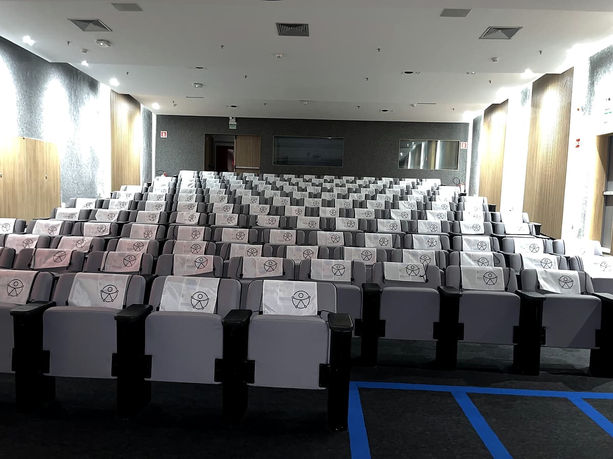 Auditório com fileiras de poltronas cinza cobertas por capas brancas com ícone de acessibilidade.