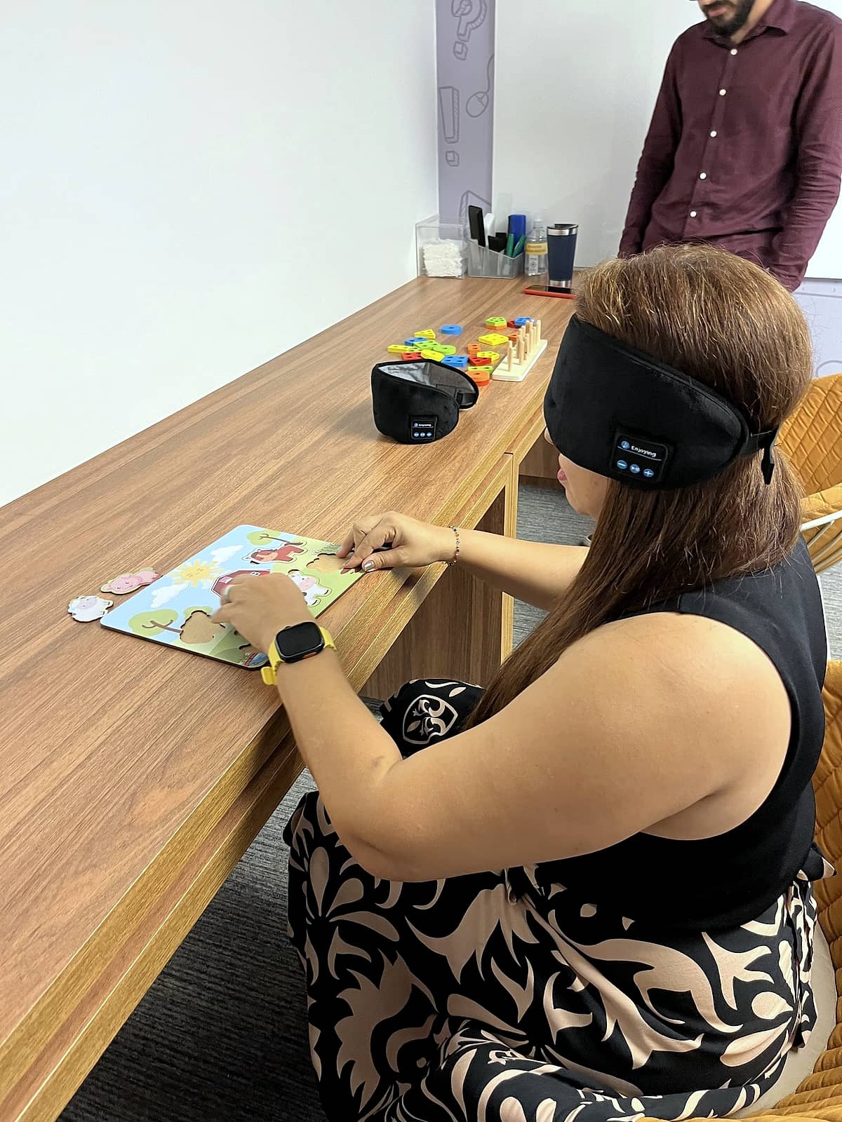Mulher com venda nos olhos montando um quebra-cabeça colorido em uma mesa de madeira; ao fundo, outro participante na mesma atividade.