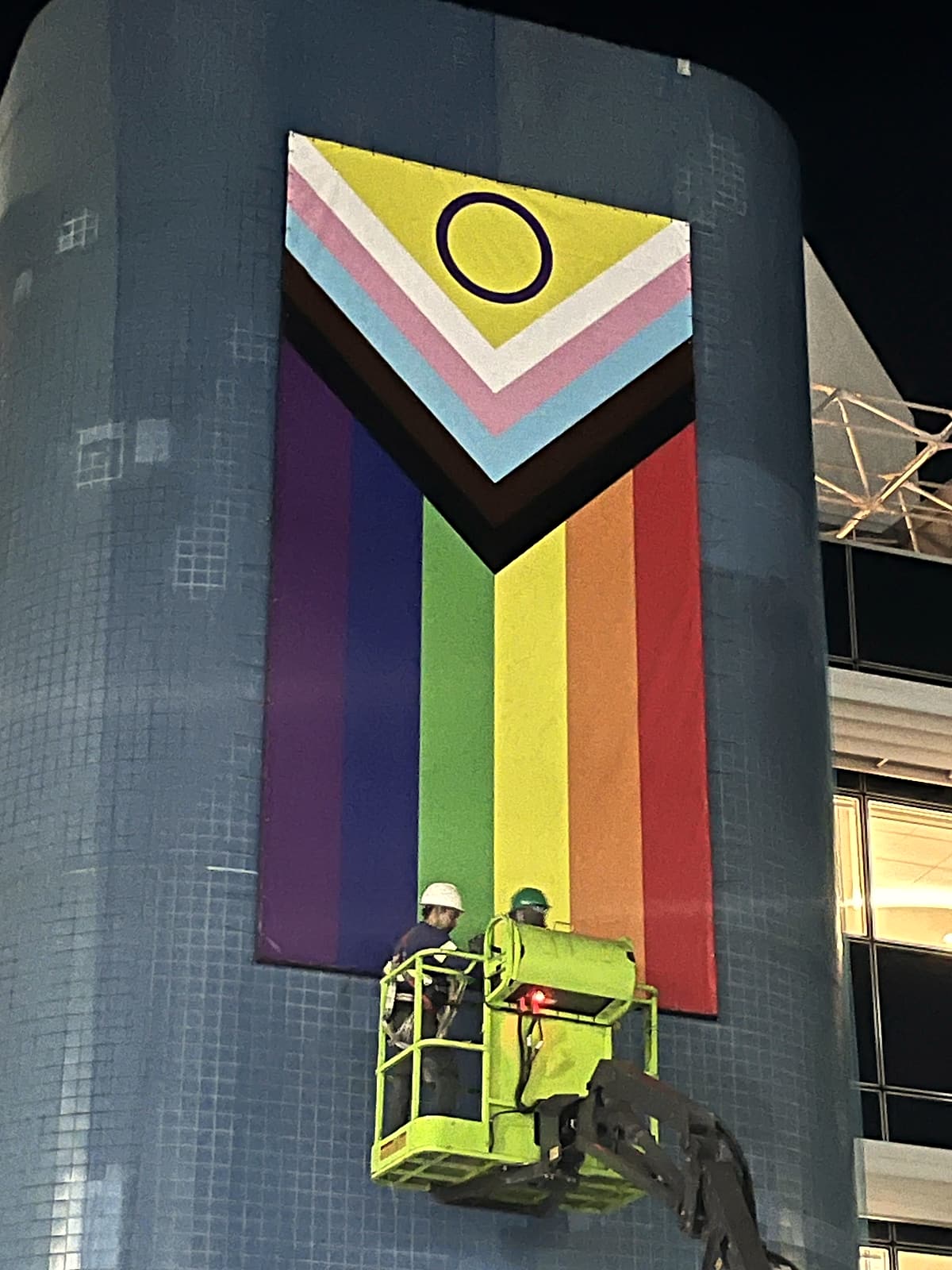 Grande bandeira do Orgulho LGBTQIA+ no formato Progress, pendurada em fachada revestida de pastilhas azuis, com dois técnicos em uma plataforma elevatória verde-limão.