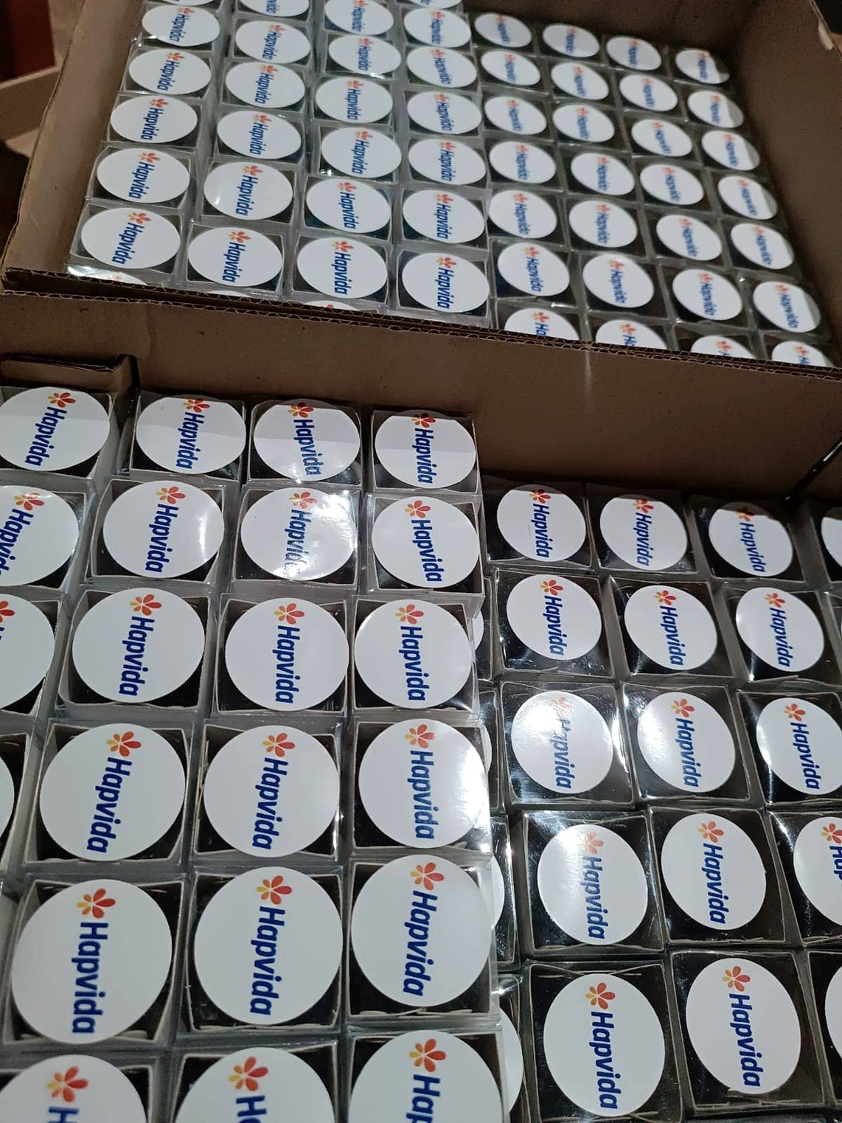 Caixas de papelão repletas de pequenos brindes brancos redondos com logo Hapvida, embalados individualmente.