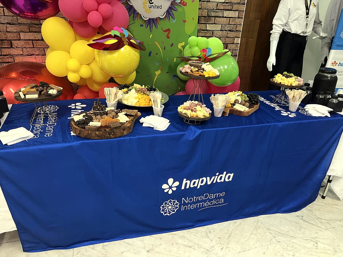 Mesa com toalha azul Hapvida NotreDame Intermédica, frios e doces, e balões coloridos com máscaras de Carnaval ao fundo.