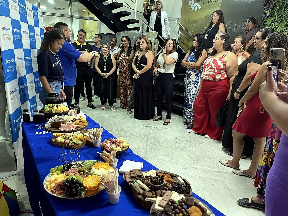 Grupo de colaboradores em pé ao redor de mesa com toalha azul Hapvida, com tábuas de frios e doces em primeiro plano.