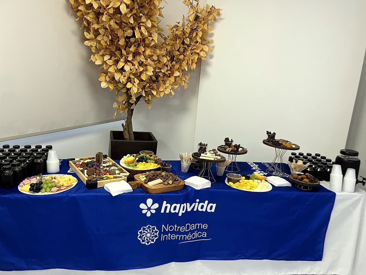 Mesa com toalha azul estampada Hapvida NotreDame Intermédica, tábuas de frios, frutas e doces, e árvore decorativa dourada.