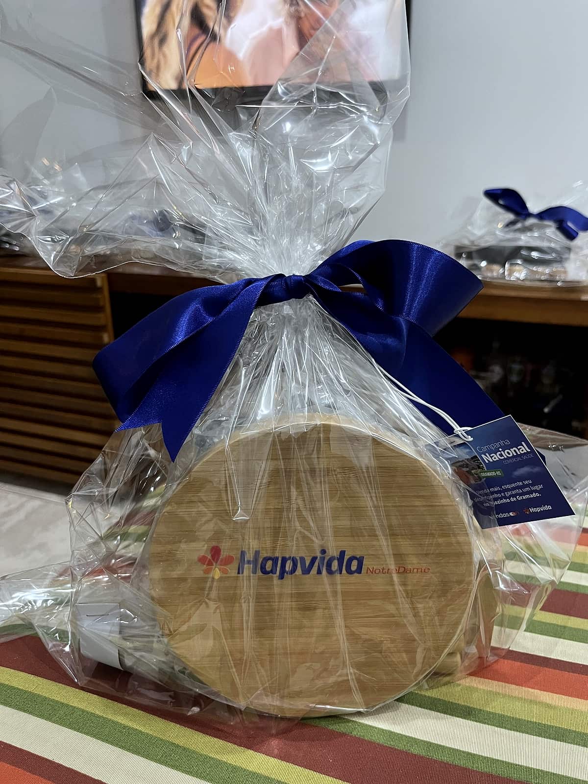 Tábua redonda de bambu gravada com logo Hapvida, embalada em celofane com laço de cetim azul e tag.