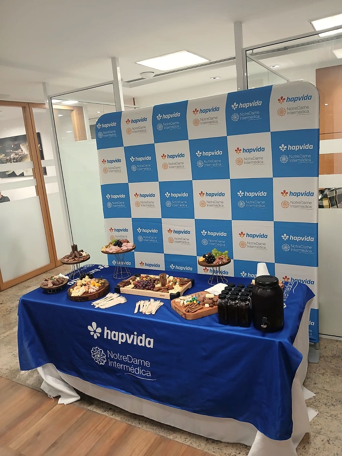 Painel quadriculado com logos Hapvida e NotreDame Intermédica atrás de mesa de frios com toalha azul.