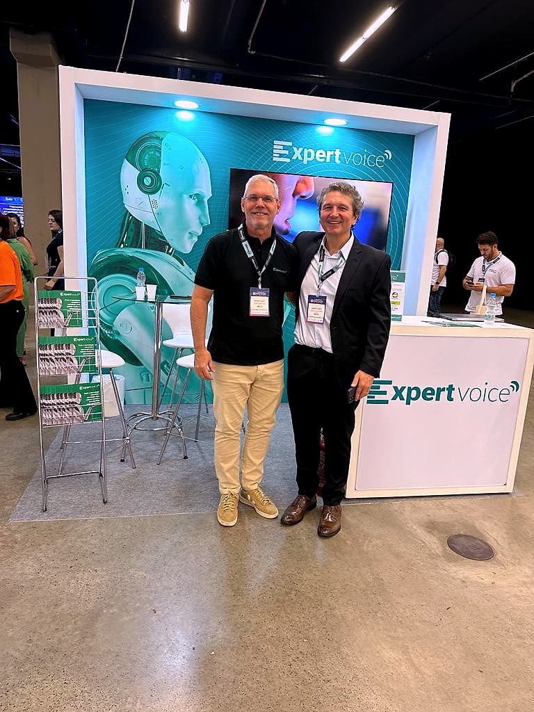Dois homens de crachá posam em frente ao estande da ExpertVoice: painel verde com um robô, balcão branco com o logo e expositor de revistas.