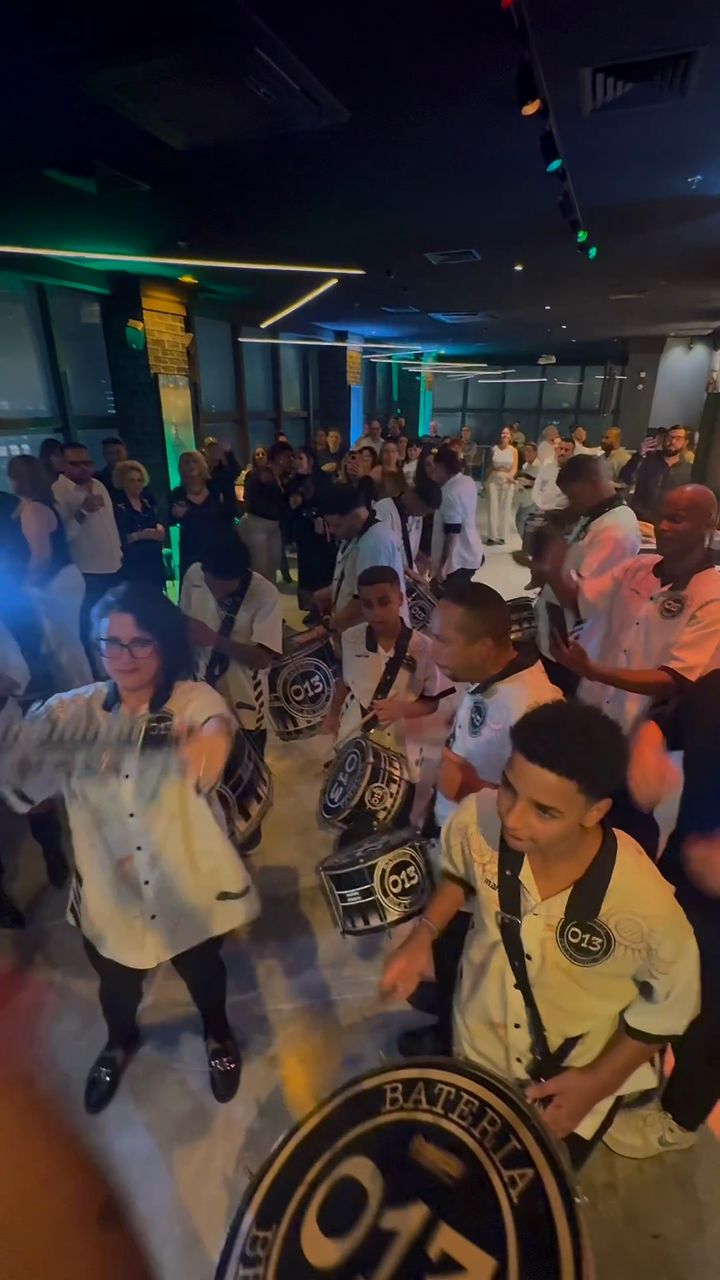 Ritmistas da Bateria 013, de camisa branca, tocam surdos e caixas no meio dos convidados em um salão com luzes verdes e azuis.