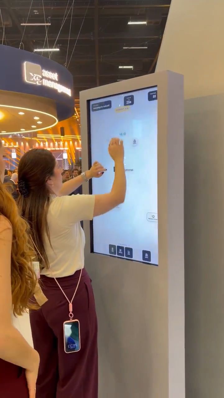 Uma participante toca uma tela touch vertical com um jogo enquanto outra aguarda ao lado, no estande.