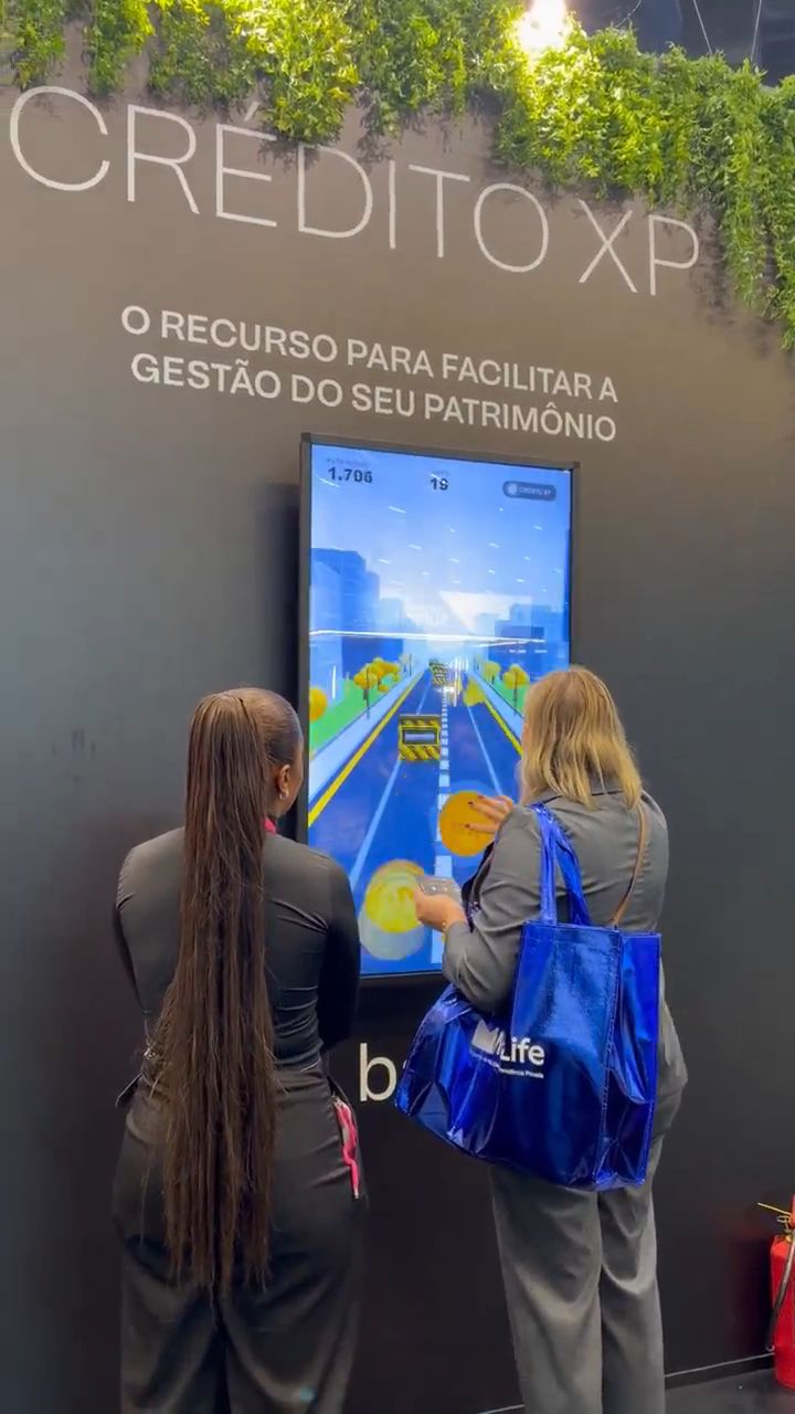 Duas participantes jogam The Yield Runner em uma tela touch vertical instalada na parede do Crédito XP.